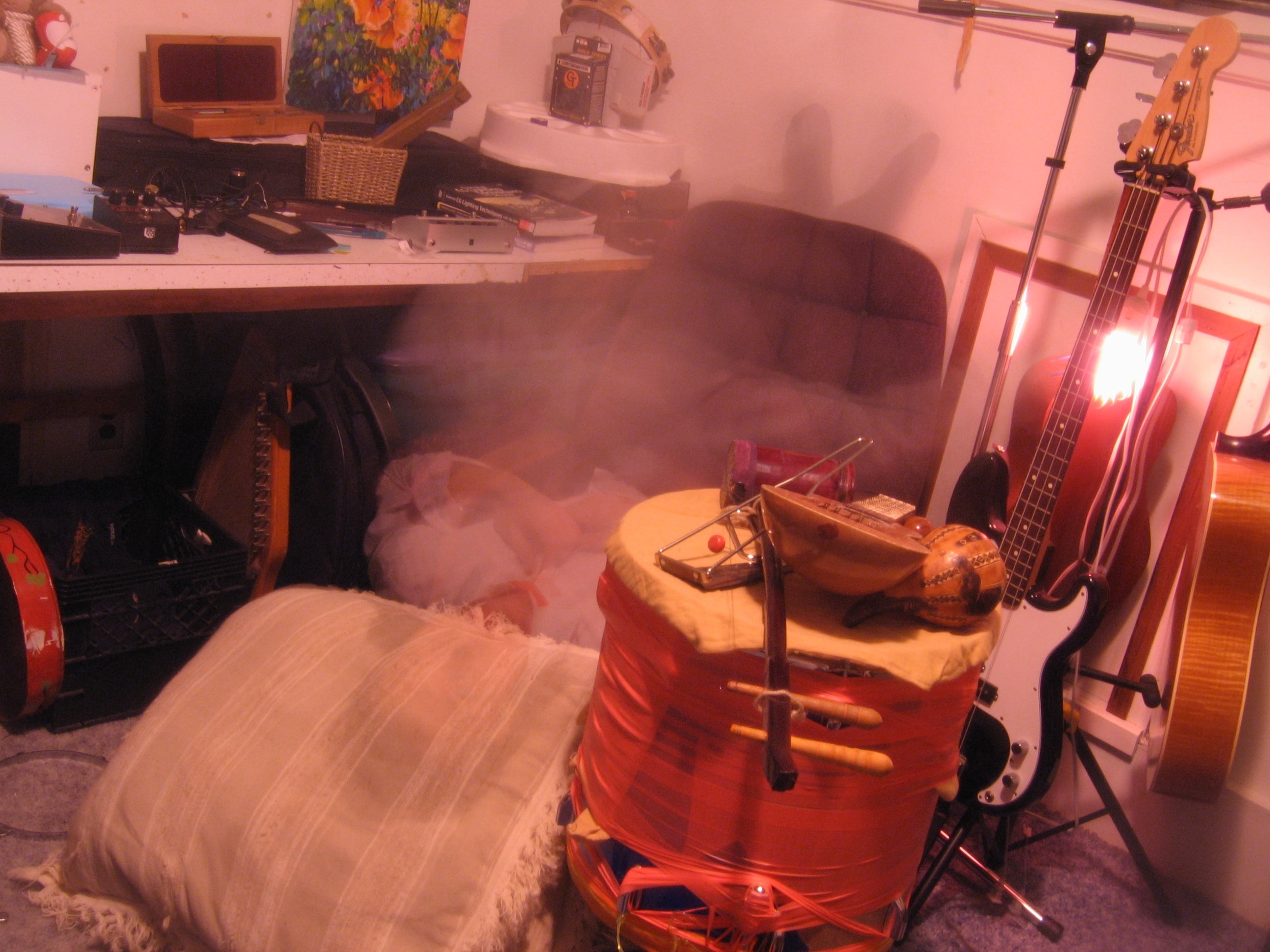 A brightly lit photo of a chair surrounded by drums, noise-makers, guitars, stompboxes, and other musical gear. In the chair, blurry from long exposure, is a ghostly figure sitting upside down — feet where their head should be, head down on a big throw pillow on the floor.