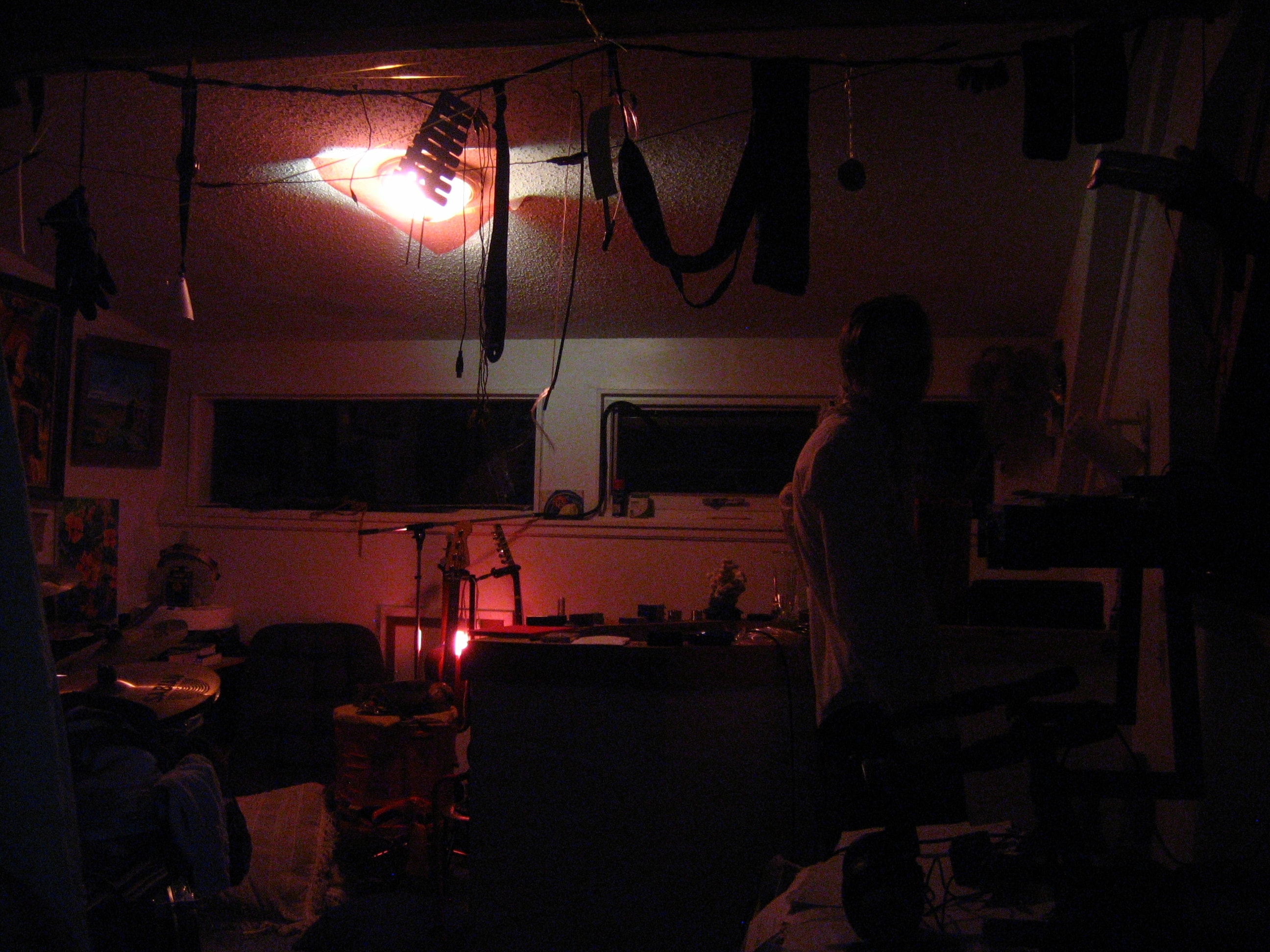 A bedroom, dimly lit in pink tones, with a barely visible person in a white shirt looking back over their slouched shoulder at the camera. The room is full of instruments, with a number of smaller ones hanging from a rope stretched across the ceiling.