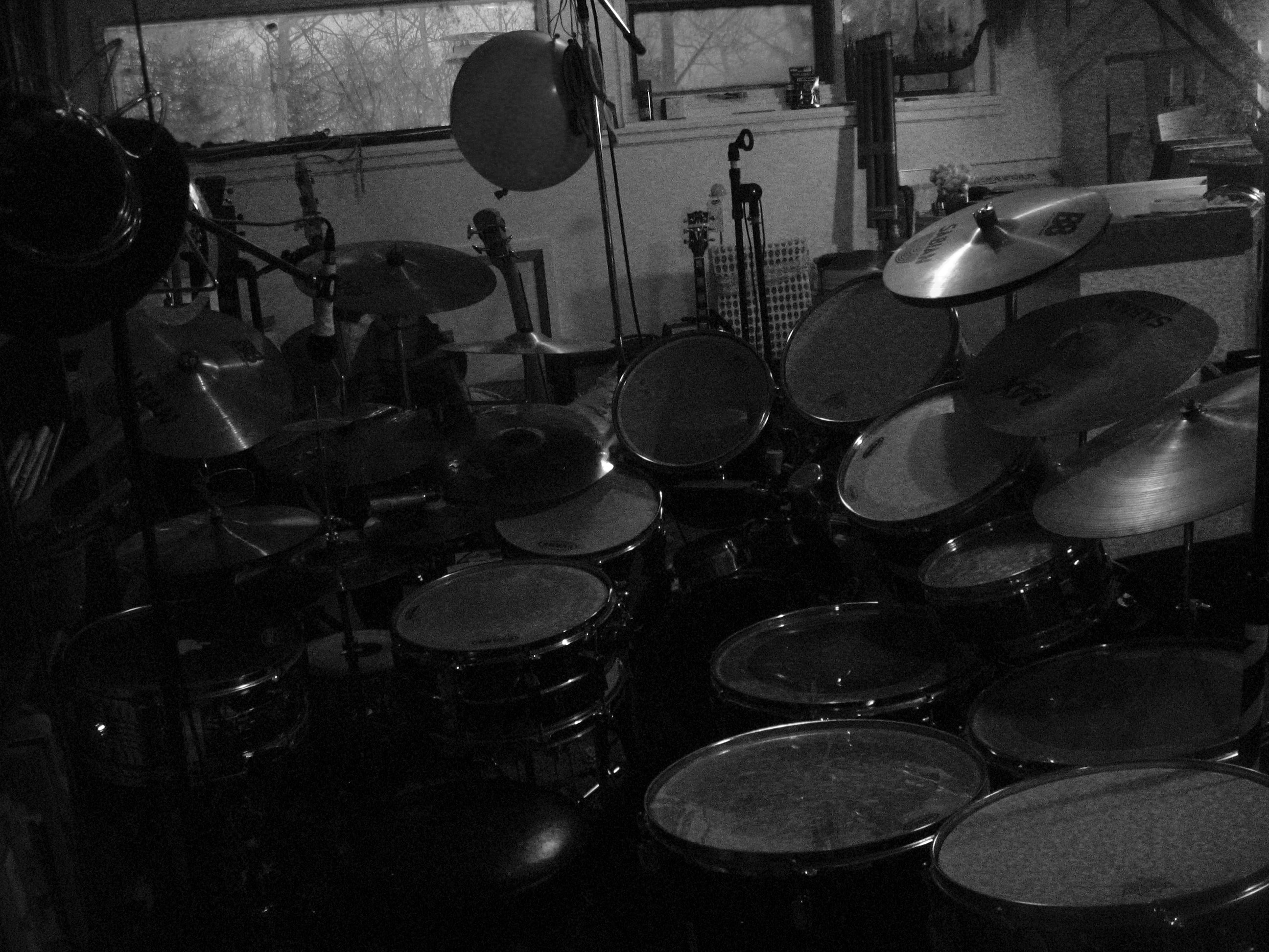 Black and white photo of a drum kit, barely illuminated. Well, that's a lie — it's three drum kits combined. Something like a dozen snares and toms, another dozen cymbals. In the background there are guitars, a very large balloon, mic stands, big long tubes that look a bit like organ pipes, and bare trees indicating late winter / early spring out the window. The point being: this is too many drums. This is a drum kit for someone who hasn't figured out how to play with tone — how to pull a good tone out of any drum — and who instead makes a lot of incoherent noise.