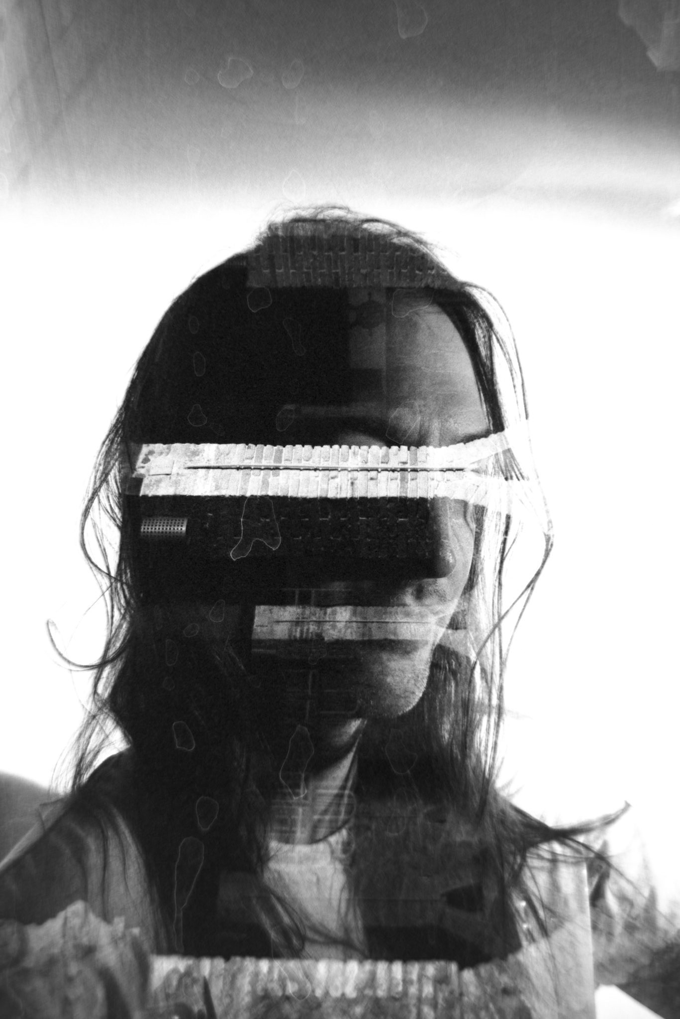 Another black and white photo. A figure with long hair that falls loosely down around their shoulders, face half in shadow, looks off to the right. Superimposed over their eyes and mouth are two bright lines, which look almost like masking tape or zippers. The photo has faint liquid stains, revealing that it was shot with film and developed by hand.