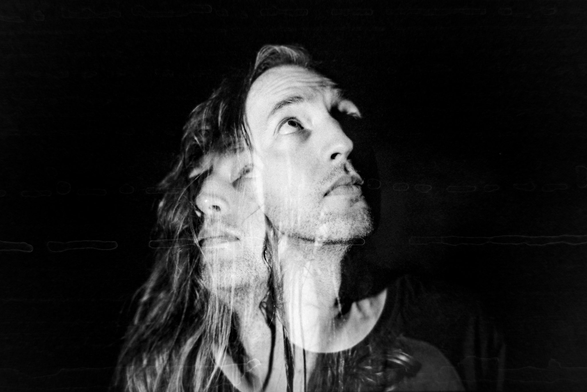 A high contrast black and white double exposure of my face, one where I'm looking up and to the right, and a second where I'm looking up to the left. The left-looking face is lower in the frame, and somewhat covered by the hair of the right-looking face. It gives the impression that there are two versions of me, one more plainly visible and one slightly hidden.