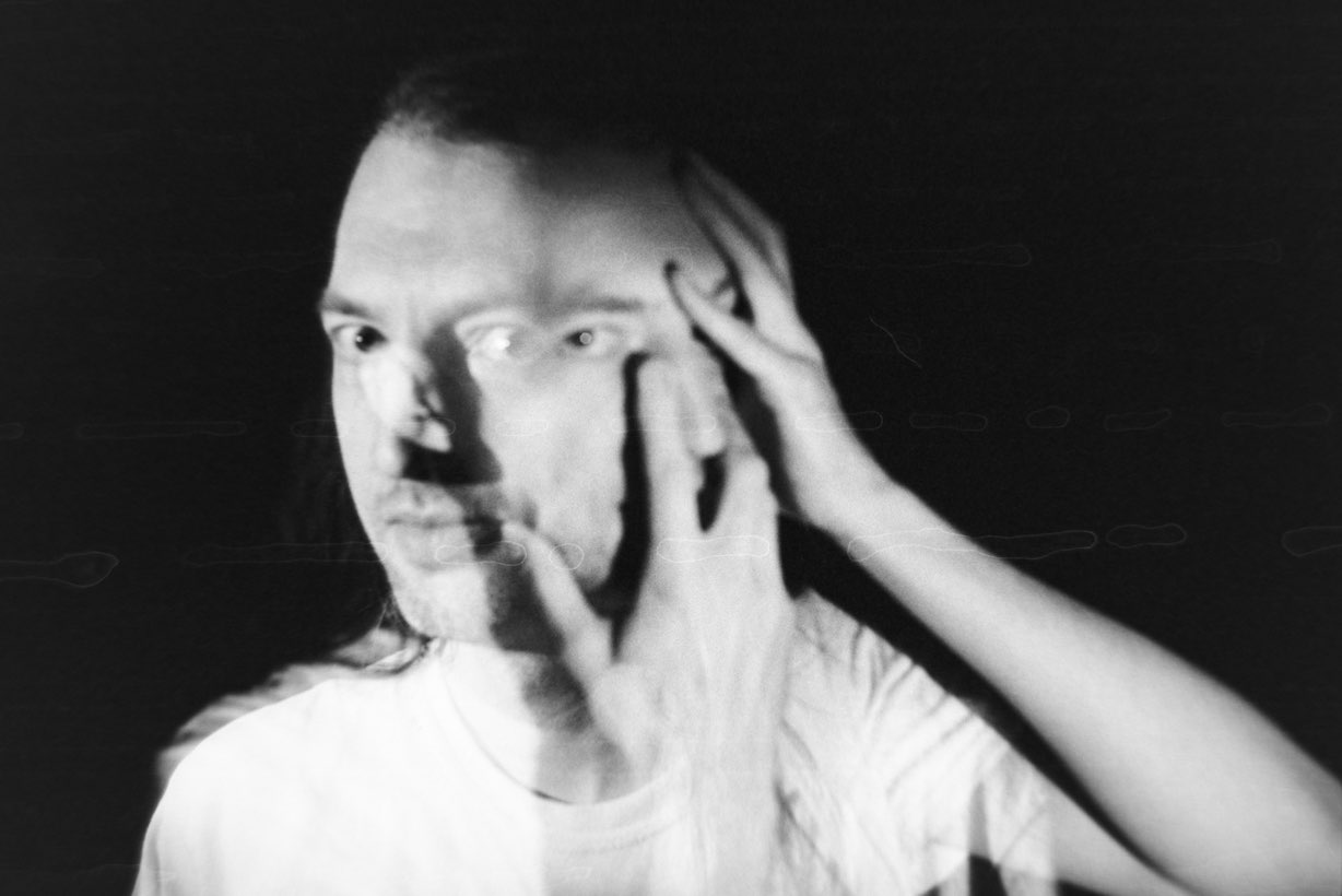 What looks like a person with two faces, shot in soft focus. One face is being covered by their hands, the other stares directly into the camera.